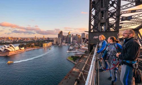 Three people standing on the Sydney Harbour Bridge looking at the harbour. Sydney Opera House and ferry in the shot.